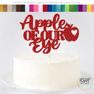 May include: A red glitter cardstock cake topper that reads "Apple of Our Eye" with a heart-shaped detail. The topper is displayed on a white frosted cake. The image also shows a variety of color options for the topper.