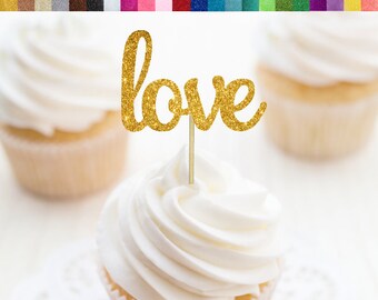 Op de afbeelding: Gouden glitter cupcake topper met het woord "love" in een cursief schrift. De topper zit op een witte cupcake met witte frosting.