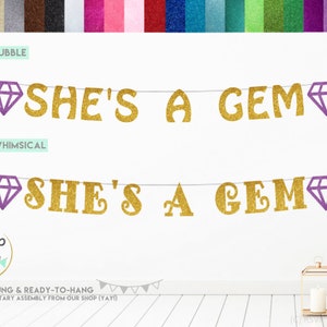 May include: Gold glitter banner with the text "She's a Gem" in two different fonts. The banner is decorated with purple diamond shapes. The banner is pre-strung and ready to hang.