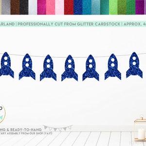 May include: A blue glitter rocket ship garland with nine rockets strung together on white string. The garland is approximately 4 feet long and is pre-strung and ready to hang. The text "RSUP Party Decor" is visible in the image.