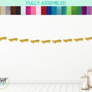 Op de afbeelding: Gouden glitter skateboard banner met 9 skateboards die aan een wit touwtje zijn geregen. De banner hangt aan een witte muur met een witte houten vloer.