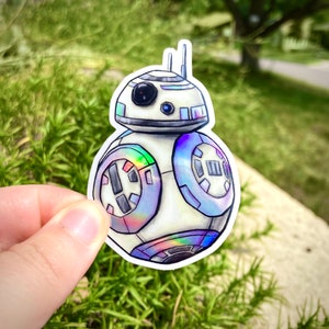 May include: A white and blue holographic sticker of a Star Wars BB-8 droid. The sticker is round and has a white background with blue details. The droid has a black eye and a white dome.