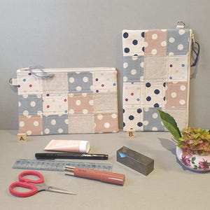 May include: Two patchwork fabric zippered pouches with a polka dot pattern in shades of pink, blue, and white. The pouches are on a light grey surface with a ruler, scissors, lipstick, and a tube of hand cream.