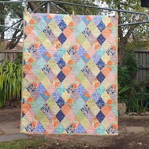 May include: A patchwork quilt with a diamond pattern, hanging on a washing line. The quilt features squares of pink, blue, yellow, and green, with floral and geometric designs. The quilt is made of fabric and is likely handmade, with a variety of colours.
