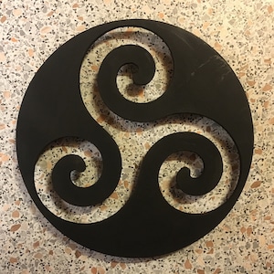 Peut inclure: Décoration murale circulaire noire représentant un Triskèle celtique. Le symbole à trois spirales est découpé dans le cercle noir plein, créant un effet d'espace négatif. Le fond est une surface mouchetée multicolore.