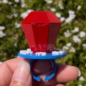 Puede incluir: Un anillo de caramelo rojo en forma de gema, con una banda azul y un borde decorativo blanco y azul. El anillo se sostiene contra un fondo verde borroso.