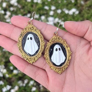 Op de afbeelding: Een paar goudkleurige, sierlijk omlijste oorbellen. Elke oorbel heeft een witte spookfiguur op een zwarte achtergrond. De oorbellen worden in een hand gehouden tegen een vage groene achtergrond.