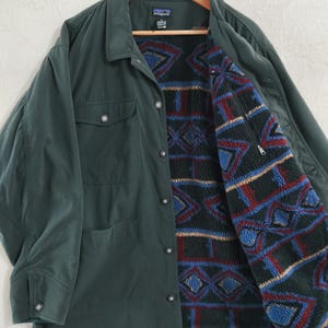 Può includere: Una giacca verde scuro con colletto, maniche lunghe e chiusura frontale con bottoni. La giacca presenta due tasche sul petto con patta abbottonata e una fodera in pile con un motivo geometrico multicolore in blu, rosso e nero.