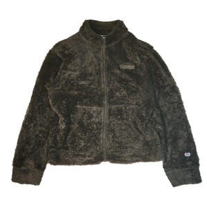 Può includere: Una giacca a maniche lunghe color marrone scuro con chiusura a zip integrale, realizzata in un materiale pile morbido e peloso. Presenta un colletto alto, due tasche anteriori e un piccolo logo Champion ricamato in rosso, bianco e blu sul polsino.