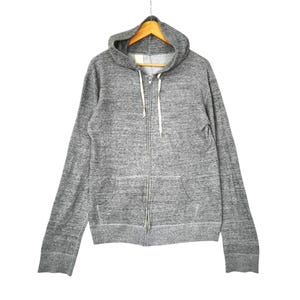 May include: A heather gray zip-up hoodie featuring a front zipper closure, a drawstring hood with white cords, and two front pockets. The garment is constructed from a textured, mottled gray fabric and is displayed on a wooden hanger.