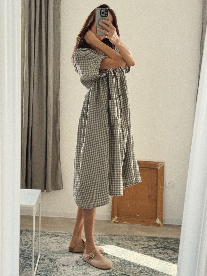 Pu&ograve; includere: Una donna indossa un abito midi a quadri vichy grigio e bianco con chiusura a bottoni sul davanti e una tasca sul lato sinistro. Indossa scarpe da barca in pelle marrone con fibbia.