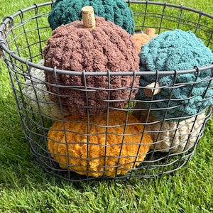 May include: A collection of six plush, crocheted pumpkins in various colors including dark green, brown, teal, mustard yellow, cream, and light green, each topped with a small cork stem, all contained within a round, dark gray wire basket.