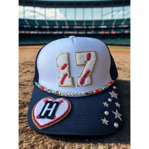 May include: A navy blue and white trucker hat with the number 17 in gold glitter and red stitching. The hat has a heart-shaped patch with the letter "H" and star and pearl embellishments.