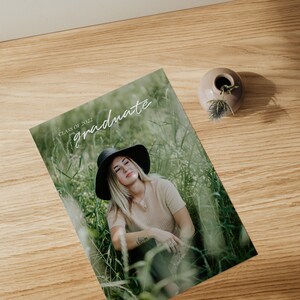 May include: A graduation announcement card with a photo of a person wearing a black hat and a beige shirt sitting in a field of tall grass. The text "Class of 2022 graduate" is written in white on the card.