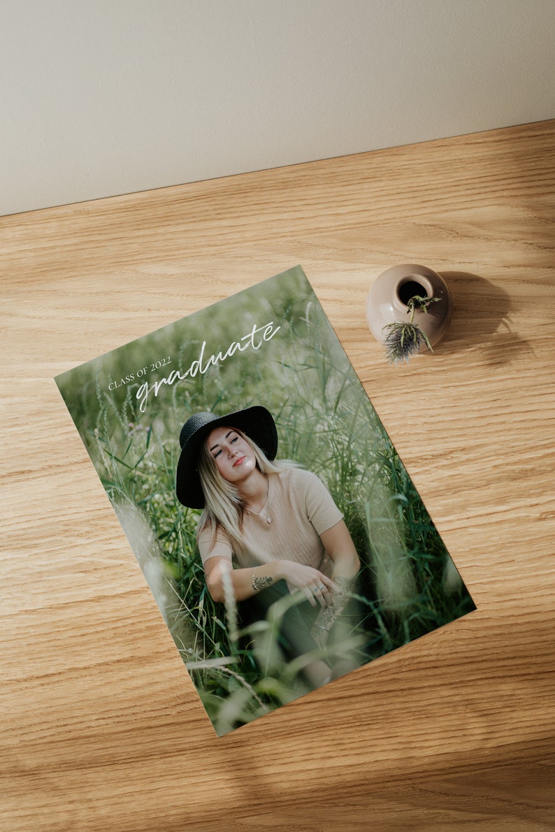 May include: A graduation announcement card with a photo of a person wearing a black hat and a beige shirt sitting in a field of tall grass. The text "Class of 2022 graduate" is written in white on the card.