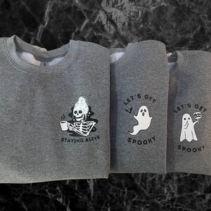 Puede incluir: Tres sudaderas grises con gráficos de temática de Halloween. La primera sudadera presenta un esqueleto bebiendo café con el texto "Staying Alive". La segunda y tercera sudaderas presentan un fantasma con el texto "Let's Get Spooky".