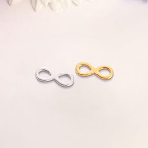 Pode incluir: Dois pingentes em forma de símbolo de infinito, um prateado e outro dourado, são exibidos sobre um fundo claro. Os pingentes são pequenos e têm um design simples. A imagem é bem iluminada, destacando os detalhes dos pingentes.