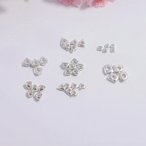 Könnte beinhalten: Eine Auswahl kleiner, silberfarbener Schmuckkomponenten in verschiedenen geometrischen Formen, darunter Dreiecke und Quadrate. Die Komponenten sind auf einer weißen Oberfläche angeordnet, mit rosa Kirschblütenzweigen im Hintergrund.