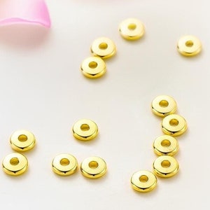 May include: A collection of gold-colored metal spacer beads, arranged in a curved line and scattered on a white surface.