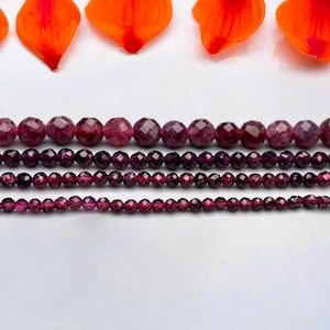 Puede incluir: Cinco hileras de cuentas de piedras preciosas facetadas de varios tamaños. Las cuentas son de color púrpura rojizo oscuro. Las cuentas están dispuestas en una fila sobre una superficie blanca.