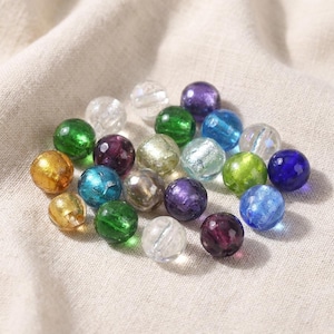 Pode incluir: Uma coleção de contas de vidro coloridas em várias tonalidades, incluindo verde, azul, roxo e dourado. As contas são redondas e transparentes, com um ligeiro brilho. Estão espalhadas sobre um tecido de cor neutra.