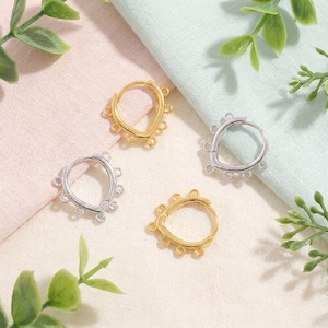 Può includere: Quattro orecchini a cerchio in oro e argento. Ogni orecchino ha una forma a goccia con piccoli anelli per attaccare ciondoli. Presentati su una superficie morbida e strutturata con fogliame verde.