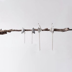 May include: Three silver butterfly earrings with dangling chains. The earrings are displayed on a branch.