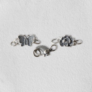 Könnte beinhalten: Drei silberfarbene Schmuckverschlüsse mit verschiedenen Designs: ein Schmetterling, ein Fisch und eine Blume.