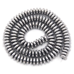 Könnte beinhalten: Eine aufgerollte Schnur aus glänzenden, silberfarbenen, flachen, runden Perlen. Die Perlen sind aufgefädelt und bilden ein spiralförmiges Muster. Die Perlen haben einen metallischen Glanz und eine glatte Oberfläche, die das Licht reflektiert. Diese Perlen sind wahrscheinlich für die Schmuckherstellung.