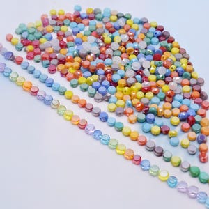May include: A collection of colorful beads in various sizes and shapes. Strands of rainbow-colored beads are arranged diagonally, with a larger pile of mixed-color beads in the background. The beads are primarily round and faceted, with a glossy finish.