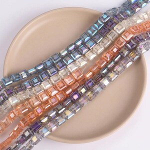 Può includere: Fili di perline quadrate e iridescenti in varie tonalità di blu, viola, arancione e trasparente. Le perline sono disposte in file su un piatto beige. Adatte per la creazione di gioielli.