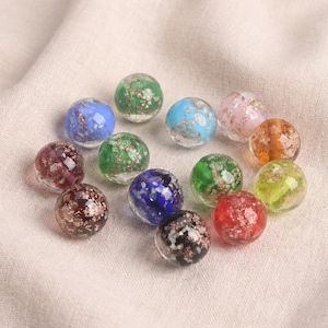 May include: A collection of colorful glass marbles. The marbles are clear with various colors, including blue, green, red, and orange, and have gold flecks inside. The marbles are arranged on a neutral-colored fabric.