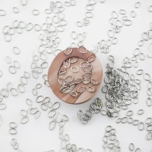 May include: A close-up of a wooden bowl filled with silver jump rings. The jump rings are scattered around the bowl on a white surface.