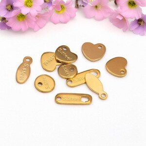 Puede incluir: Surtido de dijes de metal de color dorado con las palabras "S.Steel" grabadas en ellos. Los dijes son de varias formas, incluyendo círculos, óvalos, corazones y rectángulos.