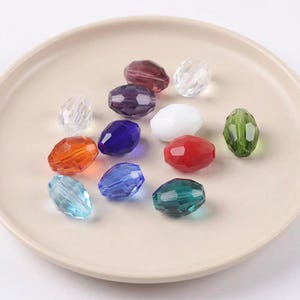 May include: A selection of faceted, oval beads in a range of colours, including clear, orange, blue, red, green, purple, and white. The beads are arranged on a light beige plate, highlighting their glossy, transparent look. Suitable for jewellery making.