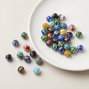 May include: A collection of colorful glass beads with various patterns, including stripes and floral designs. The beads are in shades of blue, green, red, yellow, and black. Some beads are scattered on a white plate, while others are loose on a white surface.