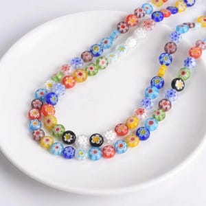 May include: Two strands of colorful millefiori glass beads. The beads are round with a floral design in various colors, including red, yellow, blue, and white. The beads are displayed on a white plate.