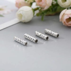 May include: Four silver-colored, geometric-patterned metal tubes. Each tube has a cylindrical shape with a faceted surface and a hollow center. The tubes are arranged on a light gray surface, with blurred flowers in the background.