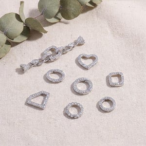 Puede incluir: Una colección de hallazgos de joyería plateados, que incluyen cierres y dijes. Los dijes tienen varias formas, como un corazón, un cuadrado y un diseño geométrico. Los cierres y dijes están adornados con pequeñas piedras transparentes.