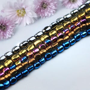 Könnte beinhalten: Eine Reihe von bunten, metallischen, facettierten Perlen. Die Perlen sind in einem abgestuften Muster angeordnet, wobei die größten Perlen oben und die kleinsten Perlen unten liegen. Die Perlen sind in verschiedenen Farben, darunter Schwarz, Gold, Blau und Lila.