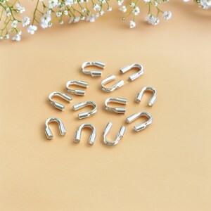 Może przedstawiać: Srebrne metalowe koraliki do zaciskania, znane również jako tulejki do zaciskania, ułożone w okrągły wzór na jasnobrązowej powierzchni. Te koraliki służą do zabezpieczania elementów biżuterii i tworzenia profesjonalnego wykończenia.