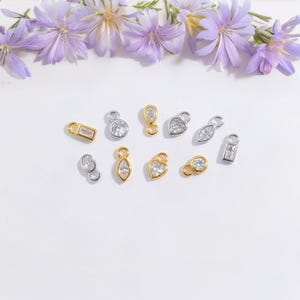 May include: A selection of small charms in gold and silver, each with a clear gemstone. The charms have various shapes, including rectangles, circles, teardrops, and hearts. The image is set against a white background, with purple flowers at the top.