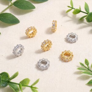 May include: An assortment of gold and silver-colored spacer beads, each adorned with clear, sparkling gemstones. The beads are arranged on a light-colored surface, with green foliage in the corners. These jewelry components are ideal for crafting bracelets and necklaces.