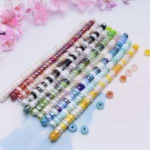 May include: A collection of colorful glass seed beads in various shades, including white, pink, red, black, green, blue, and yellow. The beads are strung together in long strands, with a few loose beads scattered nearby. Ideal for jewelry making.