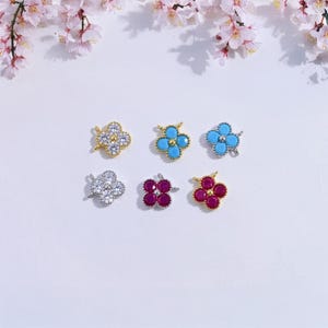 May include: Six clover-shaped charms in gold, silver, and mixed metal. The charms feature clear, turquoise, and ruby-colored stones. The charms are arranged on a white surface with pink floral accents.