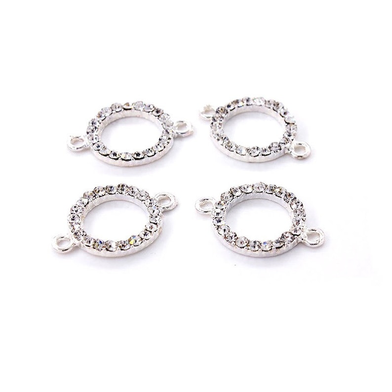 Op de afbeelding: Vier zilverkleurige metalen connector charms met heldere strasssteentjes. Elke charm heeft een rond, open cirkelontwerp met twee kleine lussen aan beide zijden.