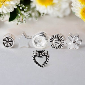 May include: A collection of silver-toned charms in various shapes, including a flower, star, thimble, and heart. The charms are detailed with embossed designs and are arranged on a white surface. The background features soft-focus flowers.