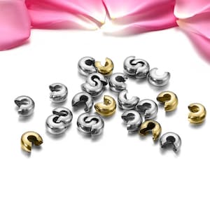 May include: A group of silver and gold metal crimp beads for jewellery making. The beads are scattered on a white surface.