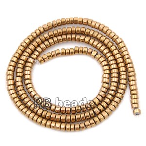 May include: A strand of gold-toned hematite beads, each bead is a small cylinder with a slightly rounded edge. The beads are strung together to form a long necklace.