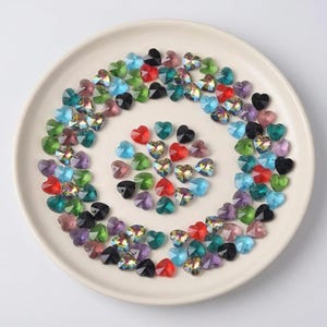 Op de afbeelding: Een verzameling hartvormige strass-steentjes in verschillende kleuren, waaronder rood, blauw, groen en paars, gerangschikt op een wit bord. De strass-steentjes hebben een facetontwerp, waardoor een sprankelend effect ontstaat. Ideaal voor knutselen en sieraden maken.